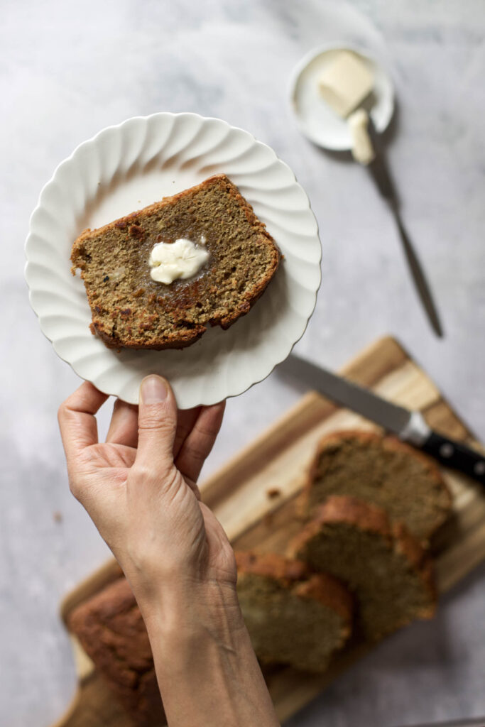 A hand holding a plate with a slice of buckwheat flour banana bread with melted butter.
